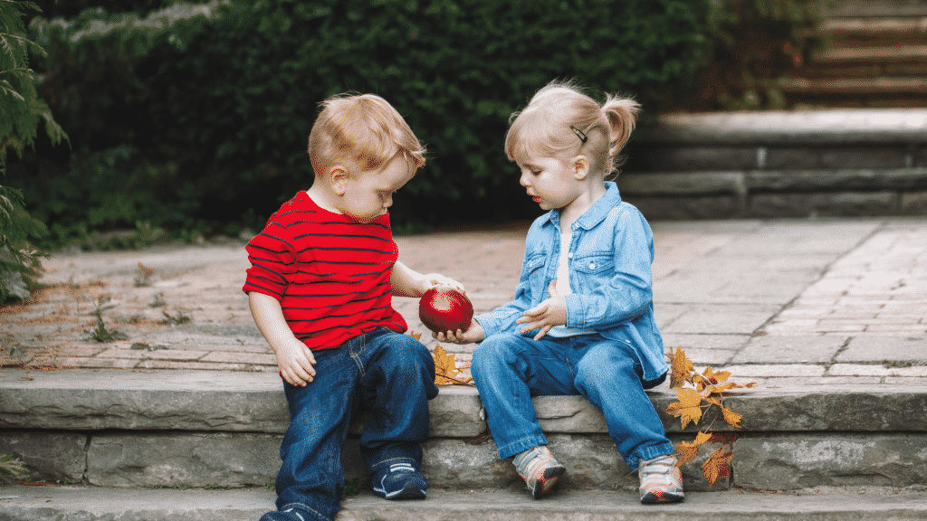 Duas crianças sentadas em uma escada de pedra sendo que a menina está dando uma maçã para o menino