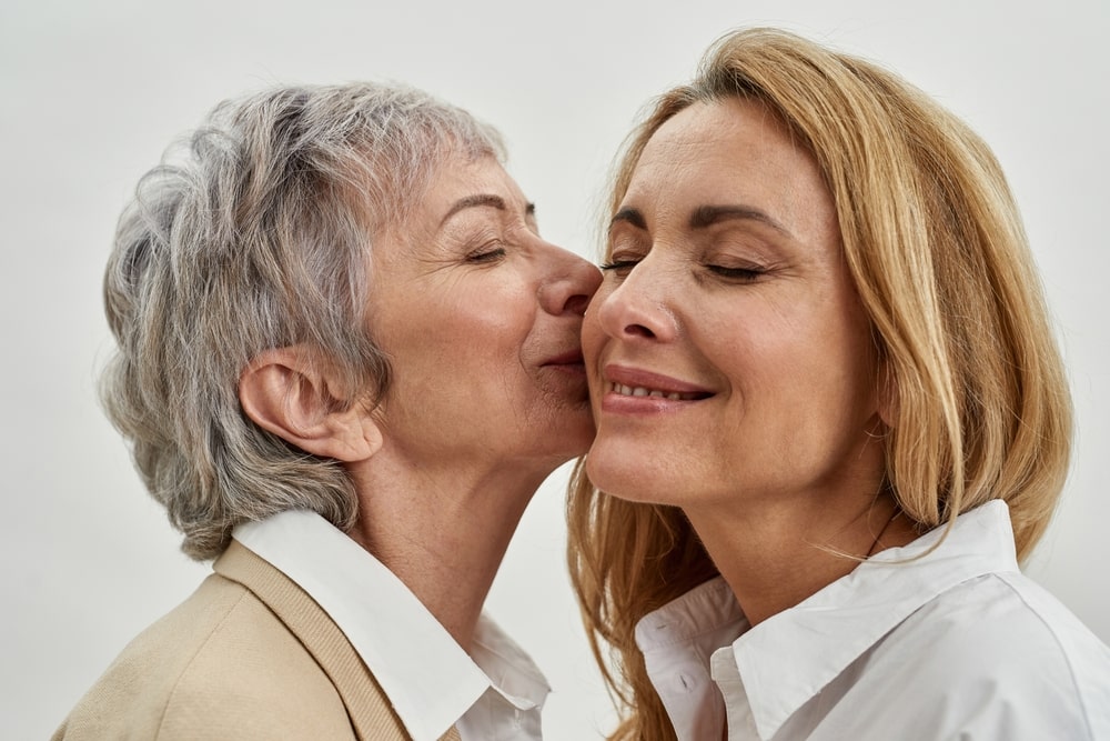 À esquerda, uma mulher branca de cabelos curtos grisalhos beijando a bochecha de outra mulher branca de cabelos loiros longos.
