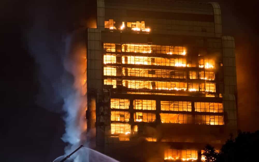Um prédio pegando fogo.  Próximo à sua parte inferior, uma mangueira de um caminhão de bombeiros.