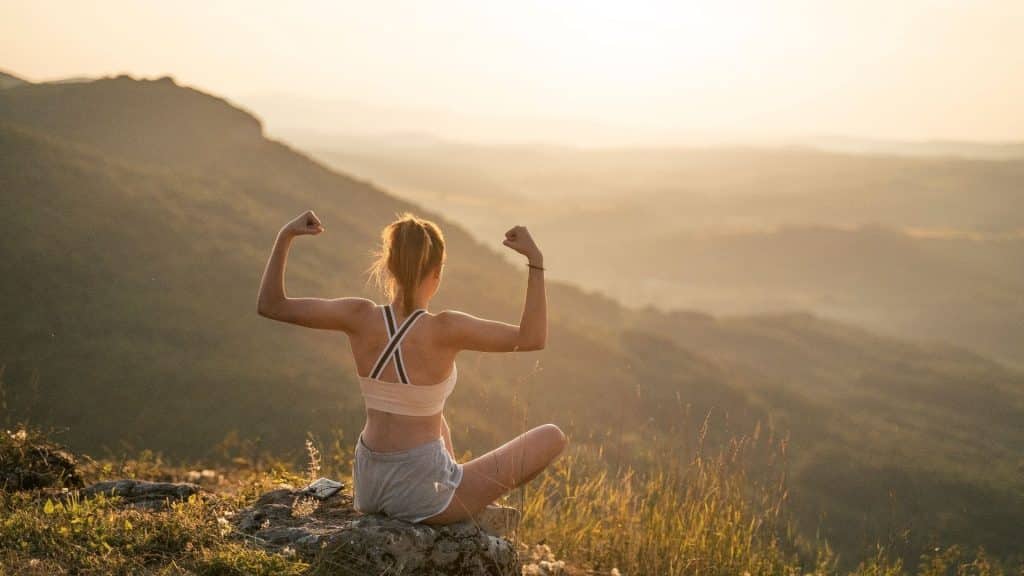 Uma mulher levantando seus braços e serrando seus punhos; gesto cuja significação é o de mostrar sua força e exibir os seus bíceps. Ao fundo, montanhas.