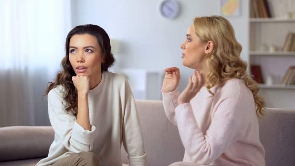 À esquerda, uma mulher aparentando descontentamento. À direita, uma mulher tentando falar com a mulher situada ao lado esquerdo.