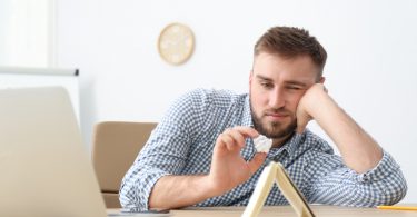 Num escritório, um homem prontificado a arremessar um papel de avião num caderno posto verticalmente numa mesa. O homem exibe um rosto entediado.