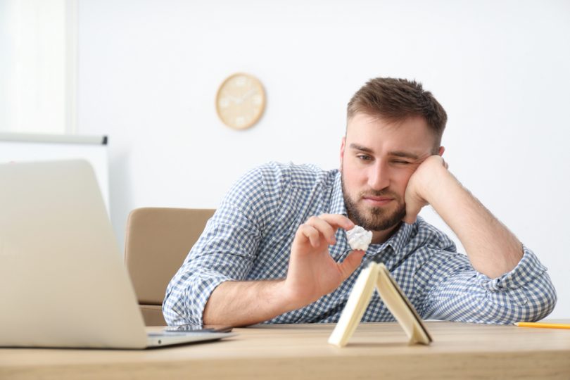 Num escritório, um homem prontificado a arremessar um papel de avião num caderno posto verticalmente numa mesa. O homem exibe um rosto entediado.