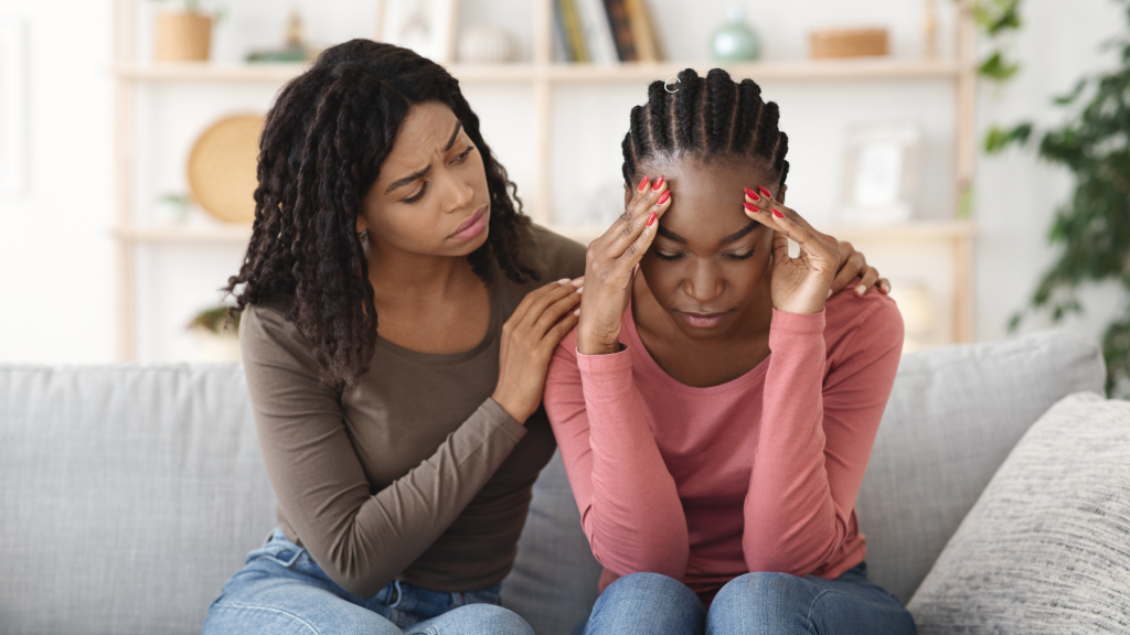 À esquerda, uma mulher consolando uma outra mulher situada ao lado direito. Ambas sentam-se num sofá.