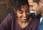 Uma mulher negra e um homem branco sorrindo. Eles tocam seus rostos um com o outro.