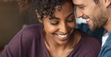 Uma mulher negra e um homem branco sorrindo. Eles tocam seus rostos um com o outro.