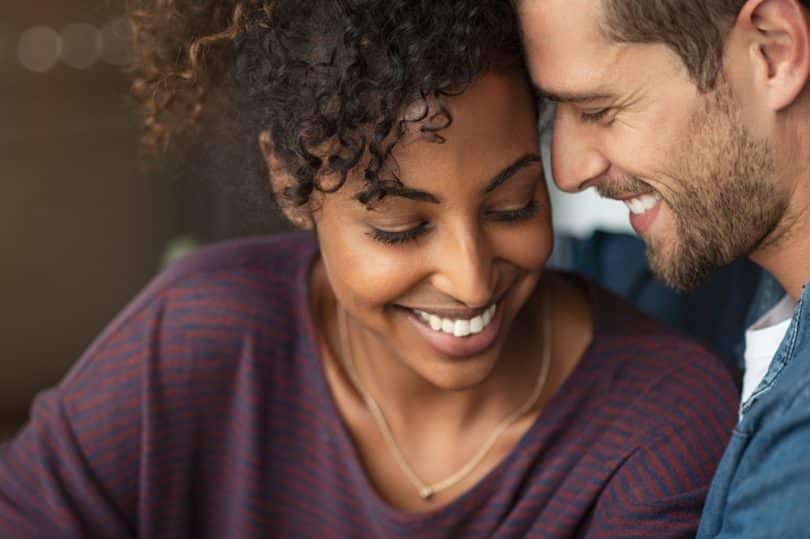 Uma mulher negra e um homem branco sorrindo. Eles tocam seus rostos um com o outro.