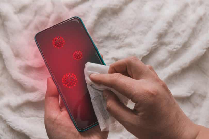 Uma pessoa limpando um celular com um pequeno pano. Sobre a tela desse celular, ilustrações que representam vírus.
