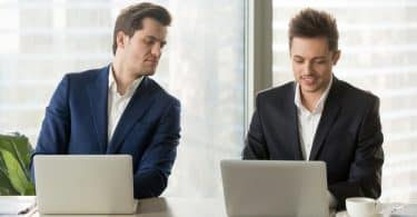 Um homem branco, à esquerda, manuseando um notebook e observando outro homem branco que também está utilizando um notebook.