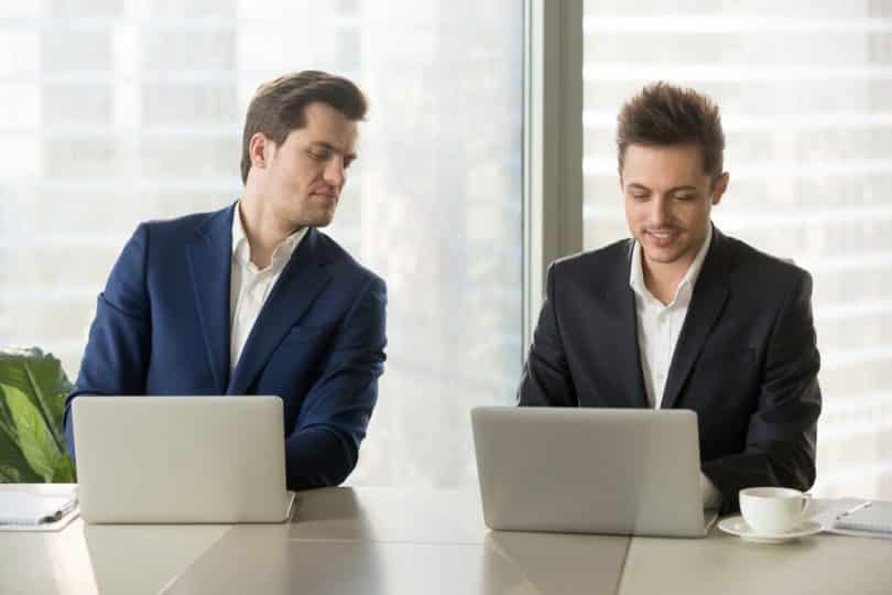 Um homem branco, à esquerda, manuseando um notebook e observando outro homem branco que também está utilizando um notebook.