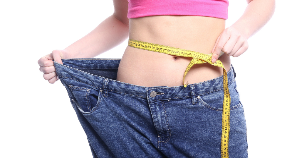 Uma mulher medindo sua região abdominal com uma fita métrica e, ao mesmo tempo, vestindo uma calça folgada.