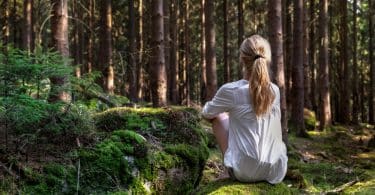 Mulher em uma floresta, sentada de costa para a câmera, ouvindo o silêncio da natureza.