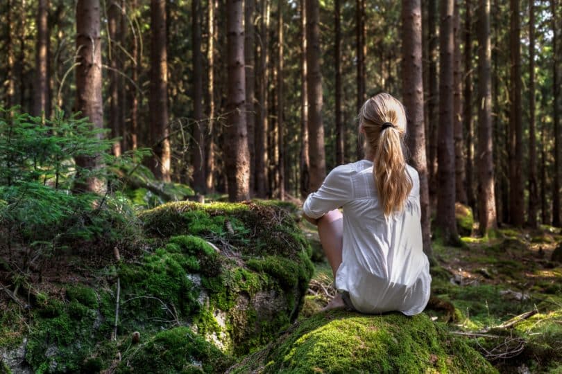 Mulher em uma floresta, sentada de costa para a câmera, ouvindo o silêncio da natureza.