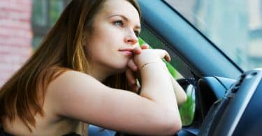 Mulher com os braços apoiados no volante do carro olhando para a frente reflexiva como queixo encostado nas mãos