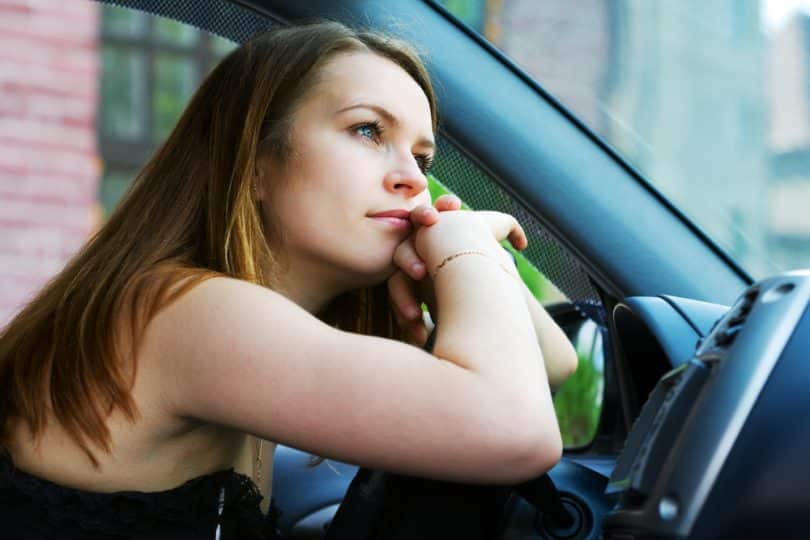 Mulher com os braços apoiados no volante do carro olhando para a frente reflexiva como queixo encostado nas mãos