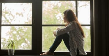 Mulher sentada em uma mureta ao lado da janela refletindo sozinha olhando para o horizonte