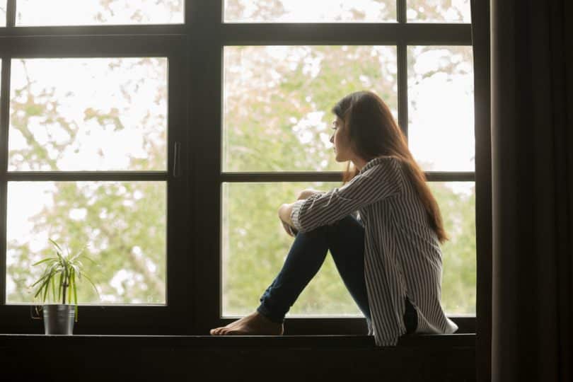Mulher sentada em uma mureta ao lado da janela refletindo sozinha olhando para o horizonte