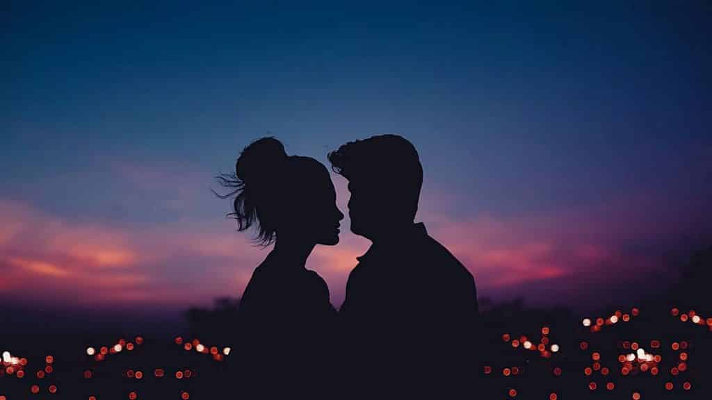 À noite, um homem e uma mulher com rostos rentes um ao outro, próximos de se beijarem. 