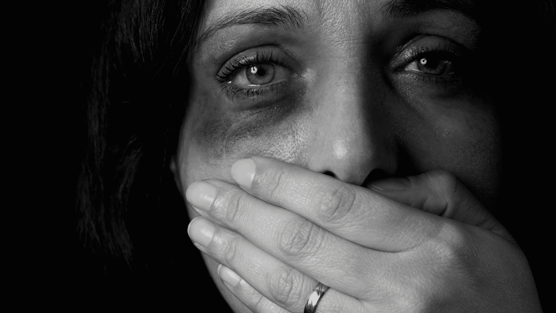 Mulher com uma mão tampando sua boca olhando para a frente com os olhos arregalados e machucados, simbolizando uma vítima de violência doméstica