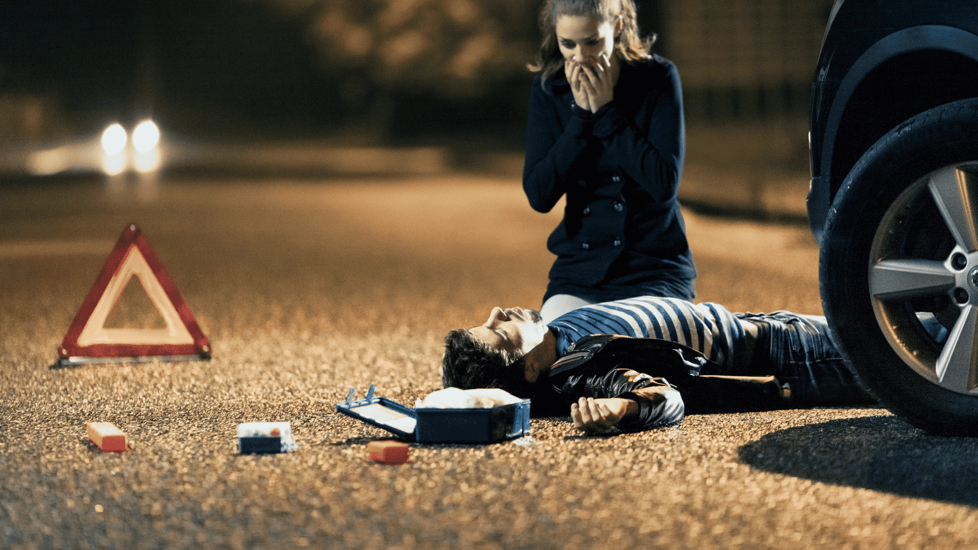 Mulher com os olhos arregalados e as mãos tampando a boca olhando um homem sem consciência no chão após um acidente de carro