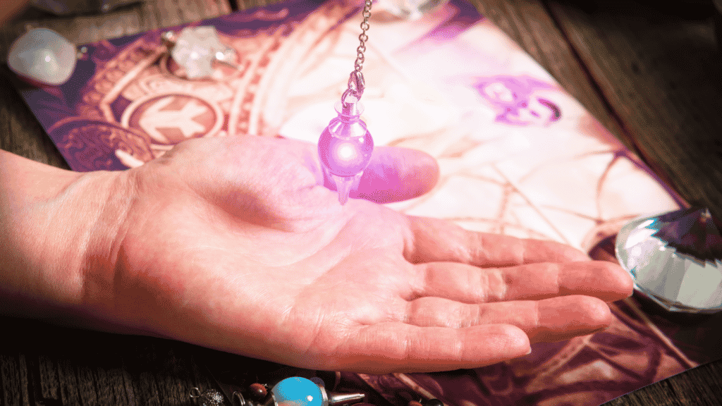 Pêndulo de cristal brilhando em cima de uma mão apoiada numa superfície com pedras de cristais e outros elementos