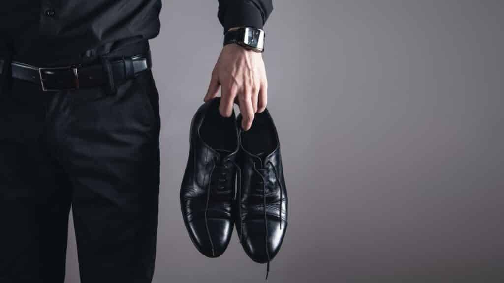 Um homem de roupas sociais segurando, com a sua mão esquerda, um par de sapatos pretos.