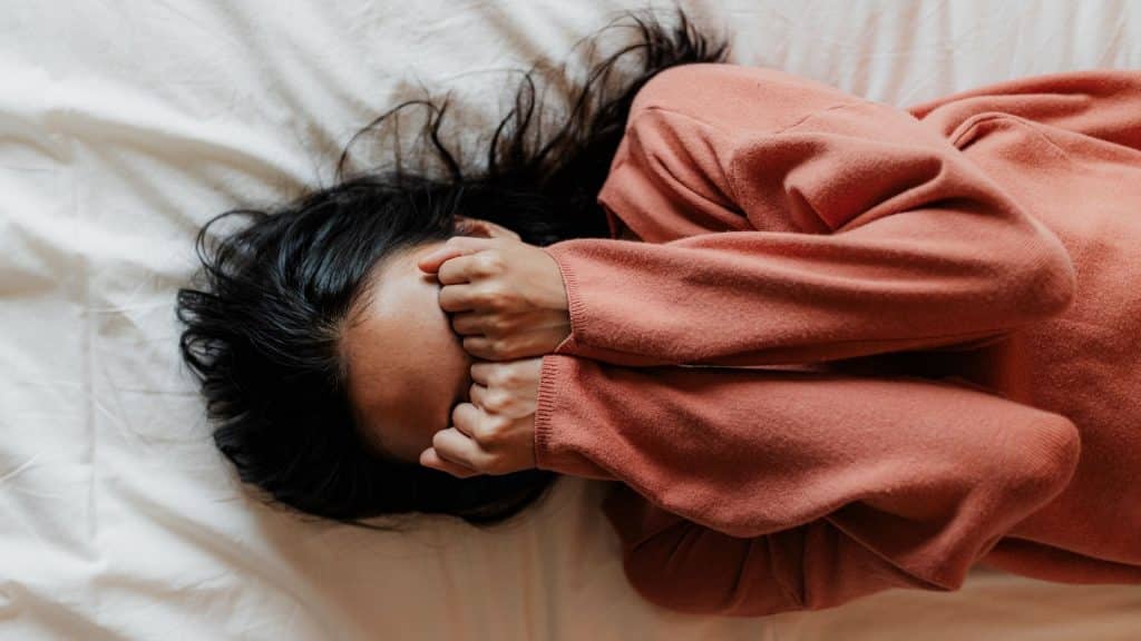 Uma mulher deitada numa cama e cobrindo o seu rosto com as duas mãos.