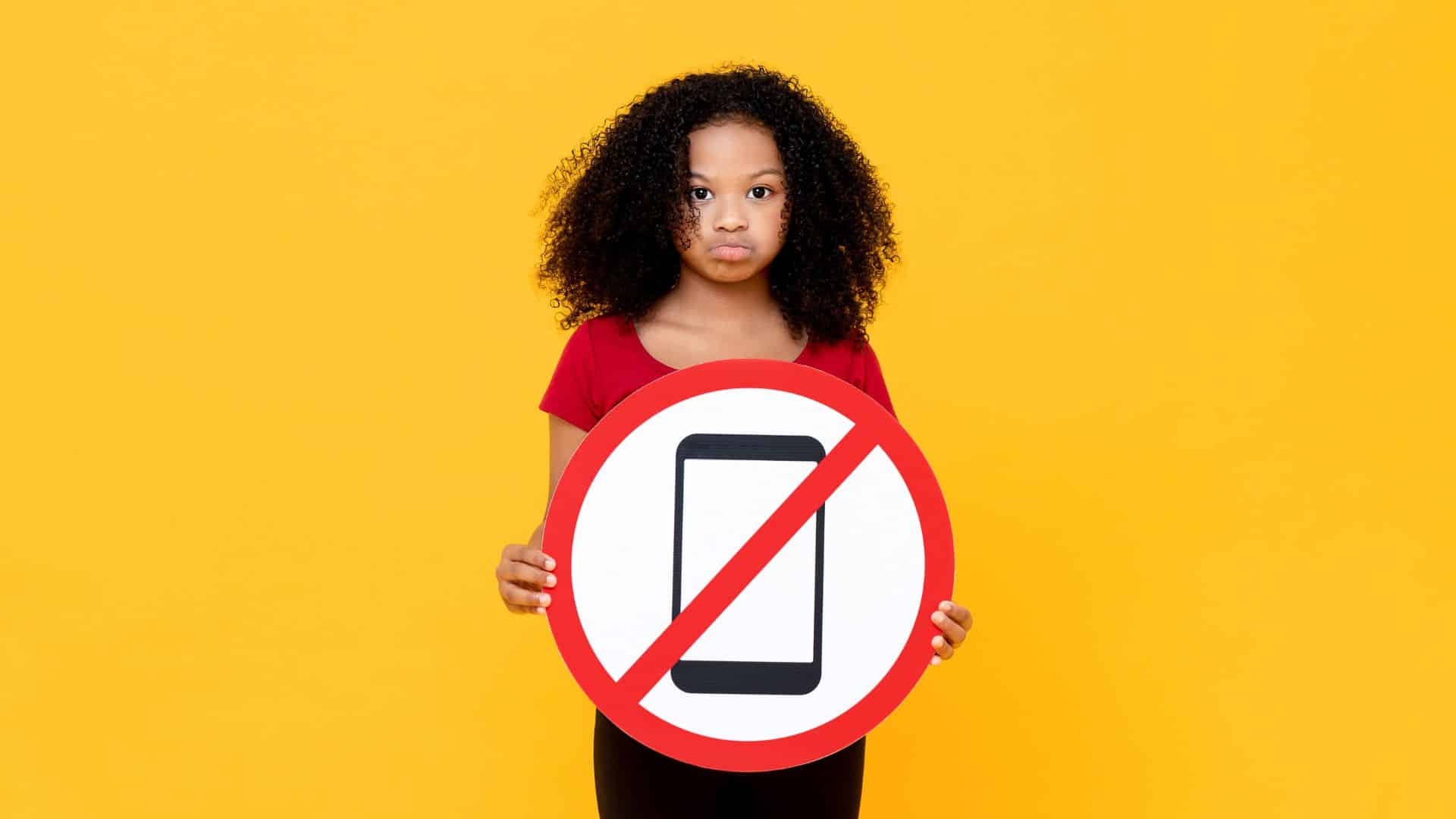 Uma mulher segurando uma placa que contém uma ilustração de um celular, e, sobrepondo-se a ela, uma faixa vermelha cuja ideia é indicar a proibição de algo. Neste caso, o celular.