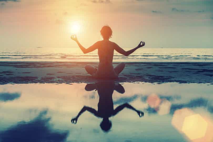 Uma pessoa meditando à beira de uma praia durante um pôr do sol.