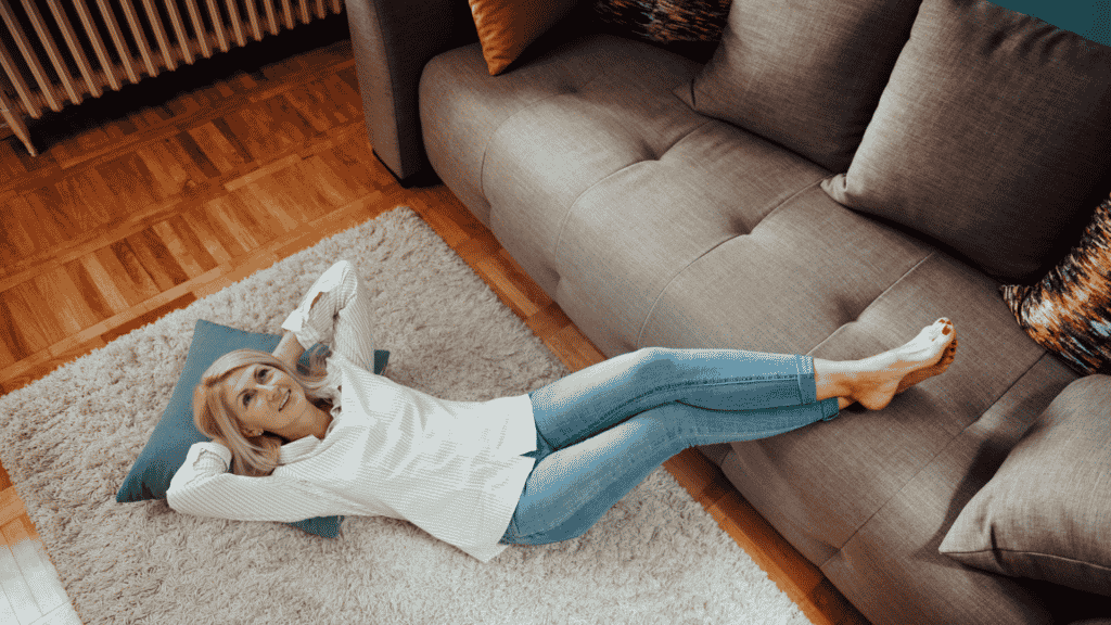 Mulher sorrindo tranquila deitada em um tapete no chão com os braços cruzados atrás da cabeça com as pernas para cima apoiadas no sofá