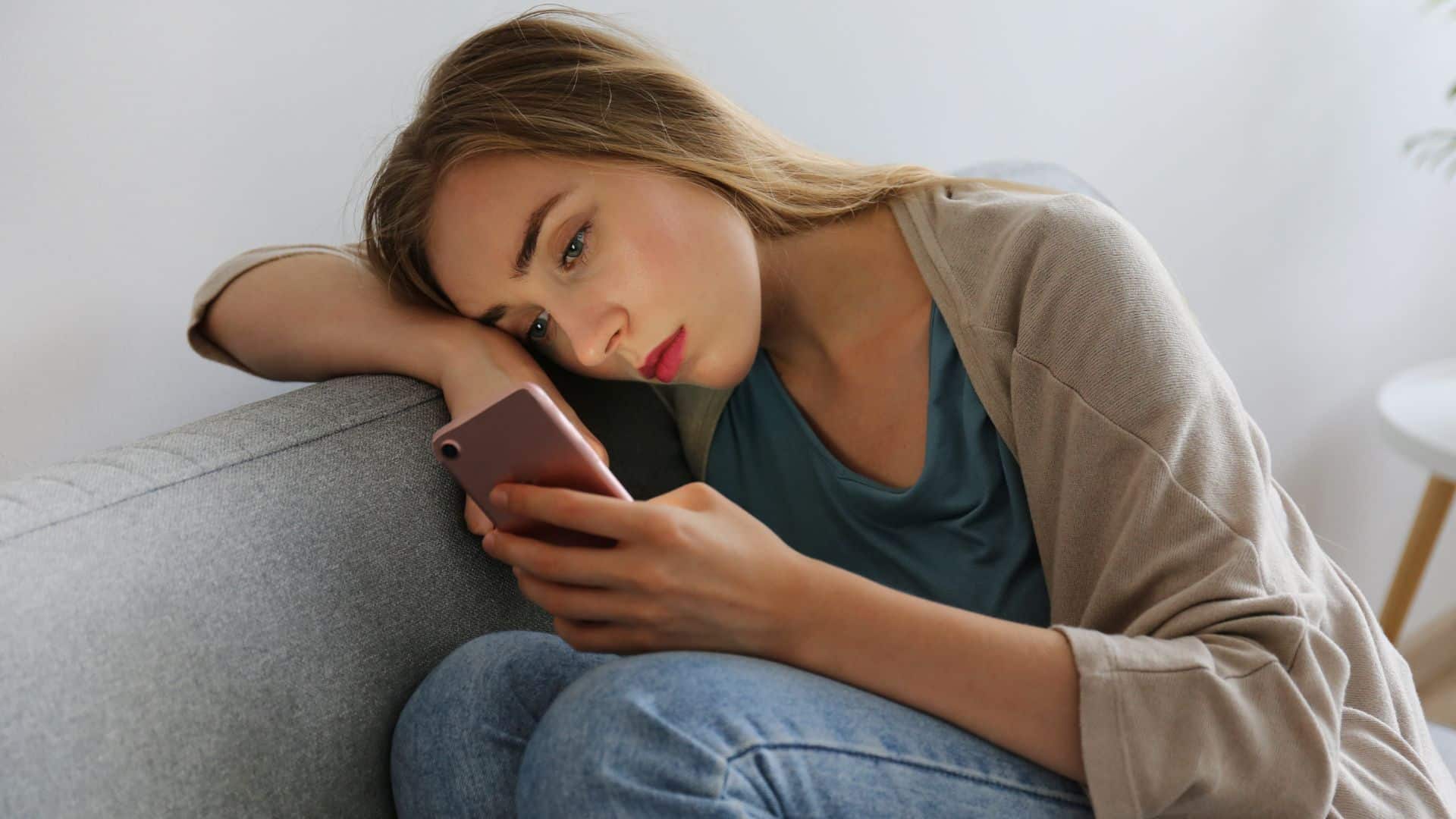Uma mulher observando a tela de um celular enquanto debruça sua cabeça na parte superior de um sofá.