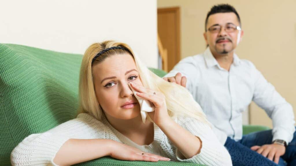 À esquerda, debruçada num sofá, uma mulher chorando. À direita, um homem tocando o ombro dela.