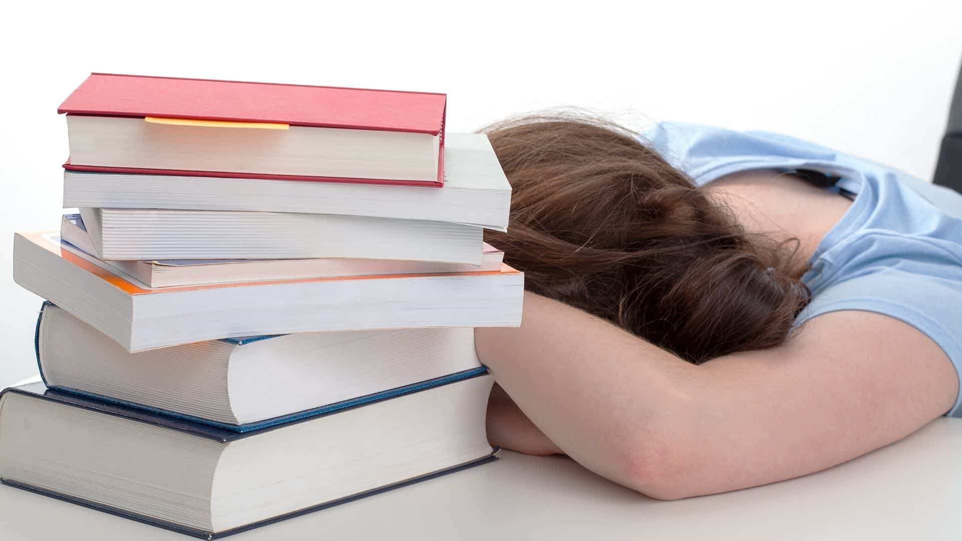 Uma criança debruçando sua cabeça numa mesa com uma pilha de livros.