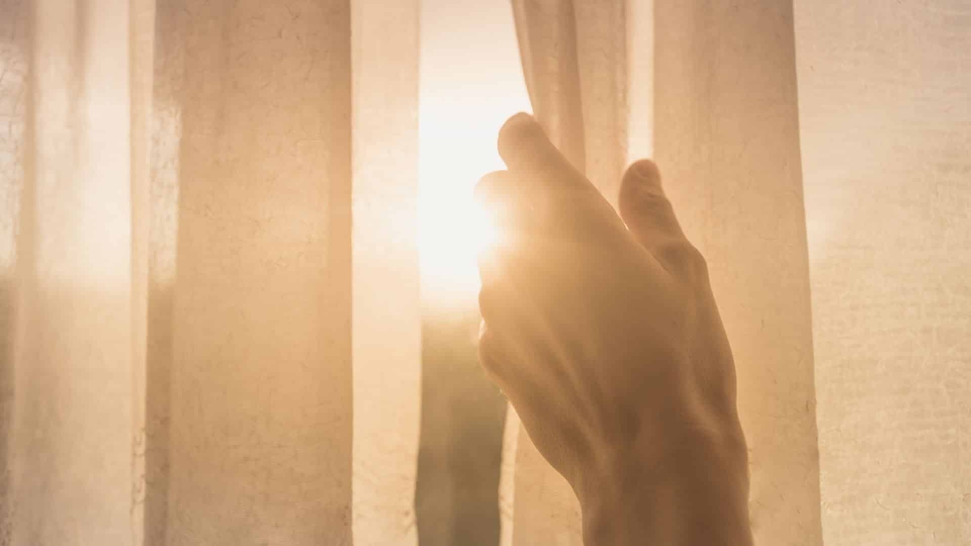 Uma mão humana abrindo uma cortina. Sua abertura revela um sol raiando.