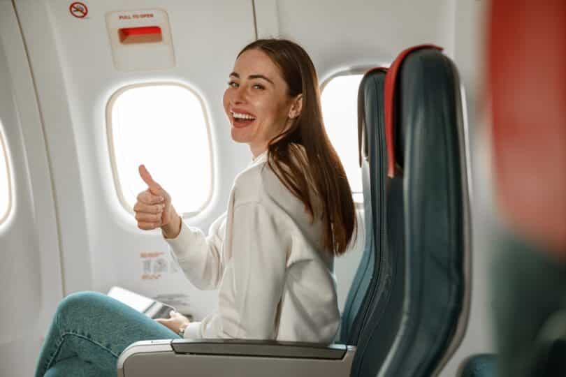 Dentro de um avião, uma mulher realizando um gesto de positivo com o dedo polegar.