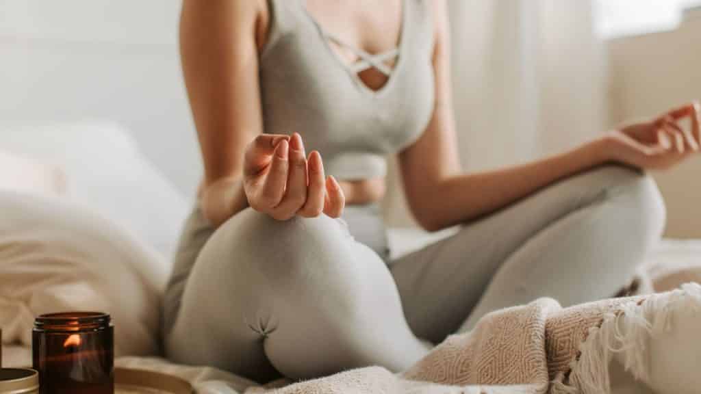 Uma mulher meditando numa cama. À esquerda dela, uma vela acesa.