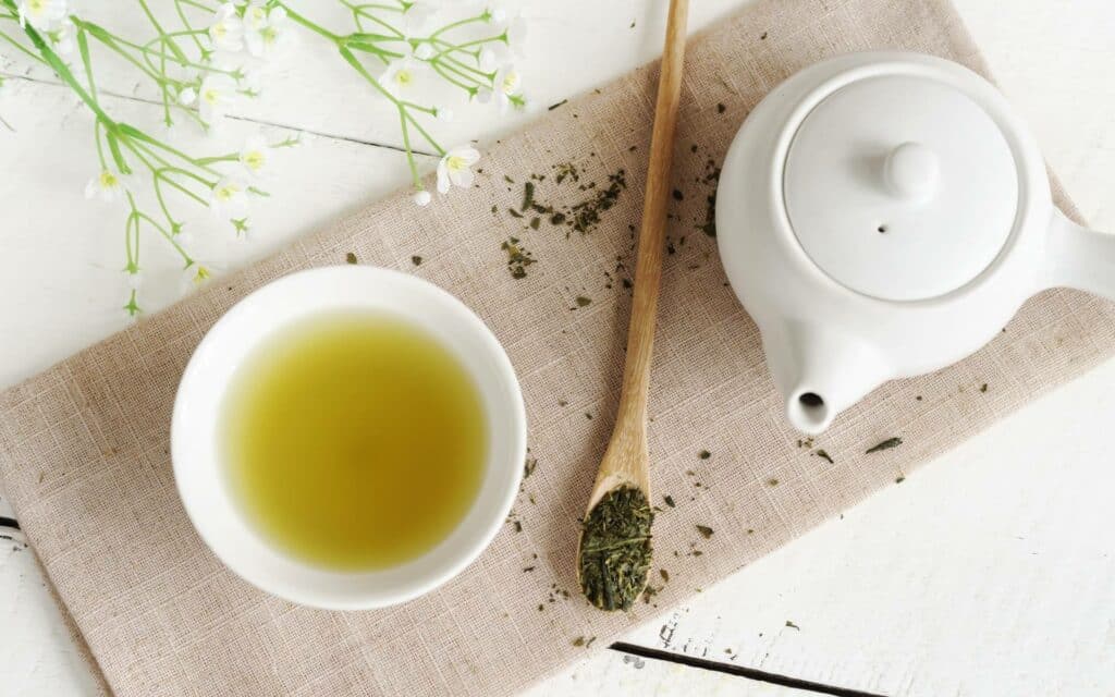 Uma xícara de chá verde disposta sobre uma tábua. À esquerda, um pequeno bule. 