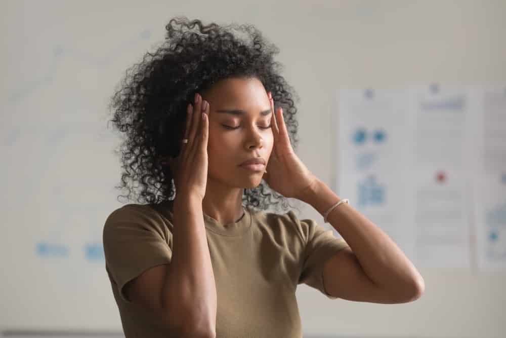 Mulher negra com os olhos fechados e as mãos na cabeça, com expressão de exaustão.