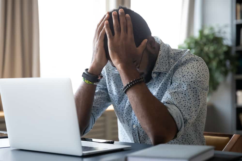 Homem negro na frente de um computador, com a cabeça baixa e as mãos na cabeça
