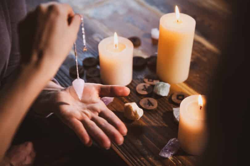 Pessoa com uma mão esticada em cima de uma mesa com velas acesas e pedras enquanto a outra segura um pêndulo de cristal