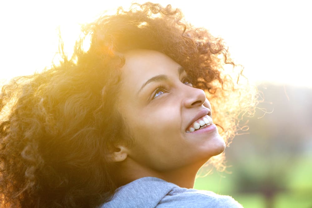 Mulher negra sorrindo e contraluz de um sol bem forte, ao ar livre