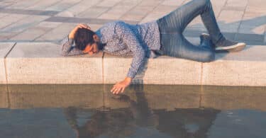 Homem deitado no chão na beira de um lago admirando para seu reflexo na água e tentando tocá-lo com a mão