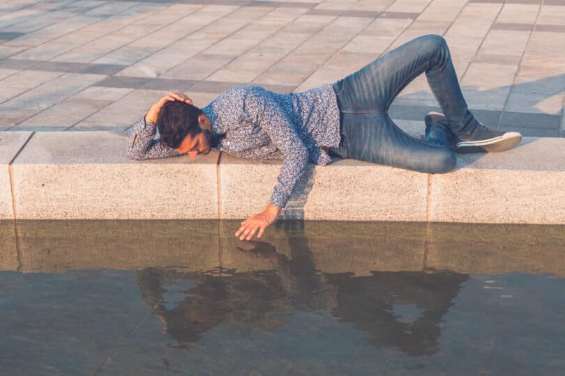 Homem deitado no chão na beira de um lago admirando para seu reflexo na água e tentando tocá-lo com a mão