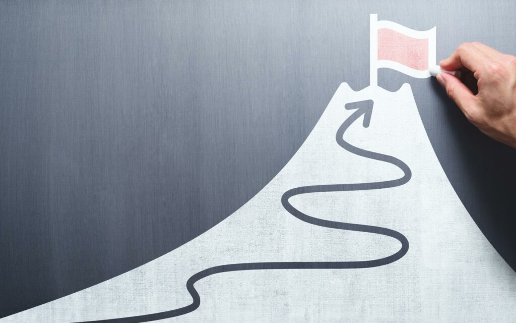 Uma ilustração de uma montanha. No pico desta, uma bandeira. Na estrutura da montanha vê-se, também, uma seta que culmina no cume da montanha.