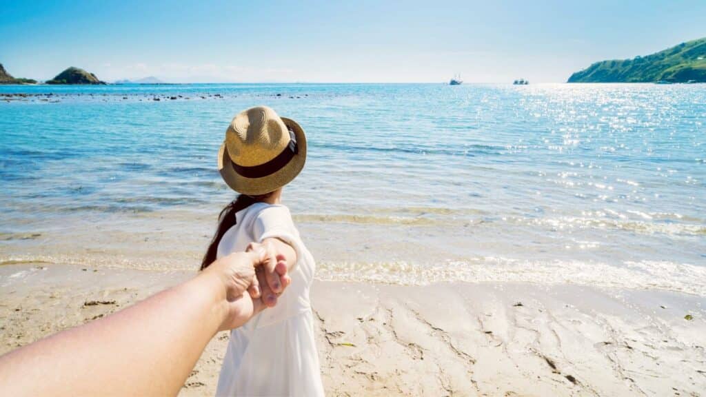 Uma mulher segurando uma mão de um homem. Ambos estão à beira de uma praia.
