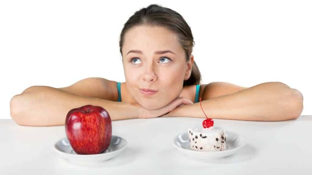 Numa mesa, um prato de maçã e um prato de bolo. Escorada sobre essa mesa, uma mulher pensativa.