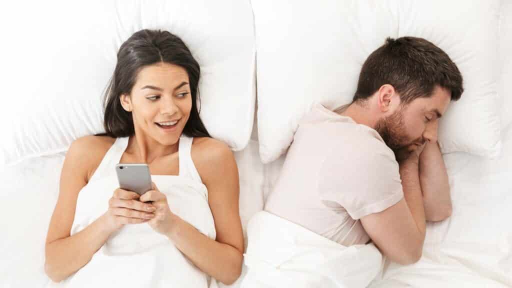 Uma mulher utilizando um celular. Ela e um homem estão deitados numa cama.