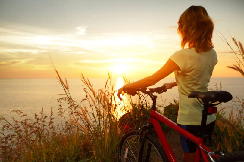 Uma mulher com uma bicicleta. Ela contempla um sol nascente.