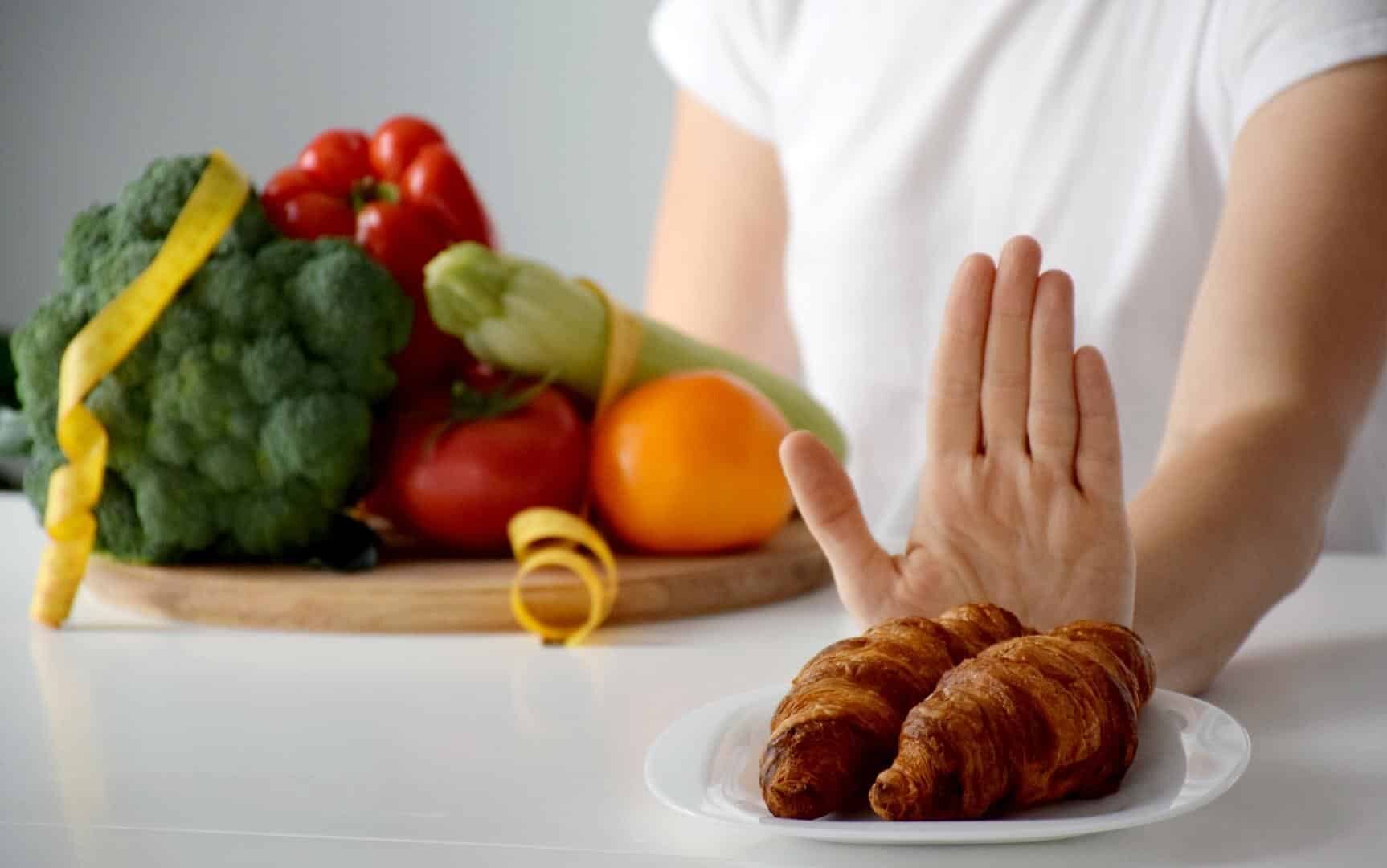 Uma pessoa afastando um prato de croissants. À esquerda, uma tábua repleta de vegetais e legumes.