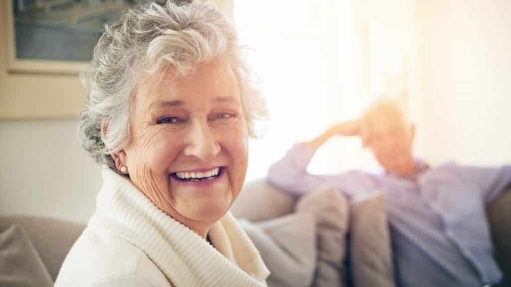 Uma mulher idosa sorrindo. Ao fundo, um homem idoso.